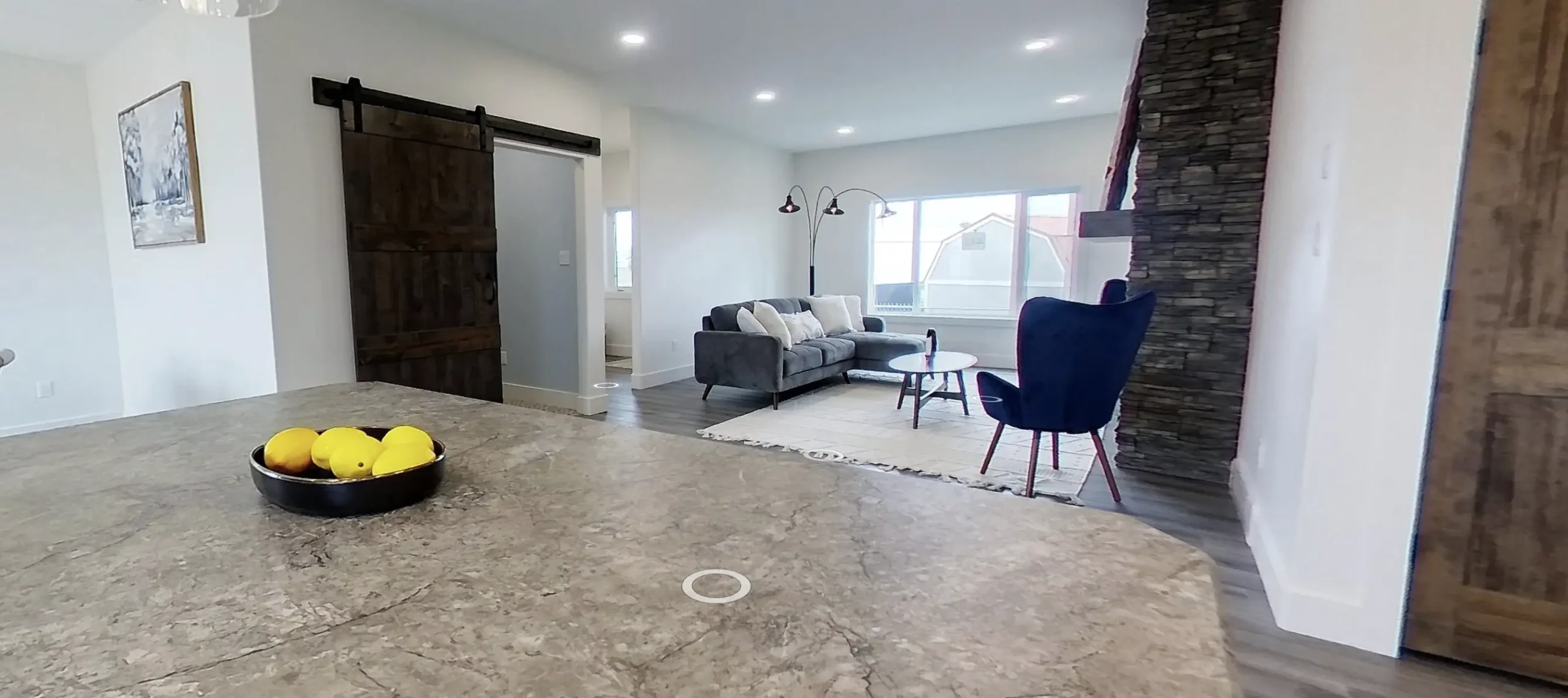RTM home in Alberta or Saskatchewan with a granite kitchen island and bowl of lemons in the foreground, rustic barn door, stacked stone fireplace, gray sofa, and blue accent chair.